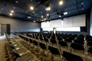 Moderne conferentieruimte aan de Johan Huizingalaan met rijen zwarte stoelen en grote projectieschermen.